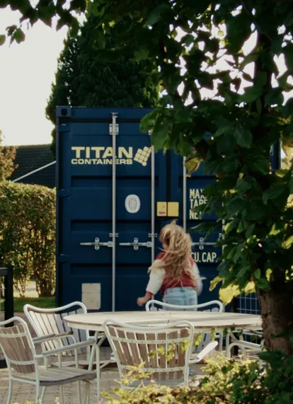 Outdoor seating area with tables and chairs in front of a blue TITAN Containers shipping container, surrounded by greenery. The container is used as a functional storage or site solution in a garden-like setting.